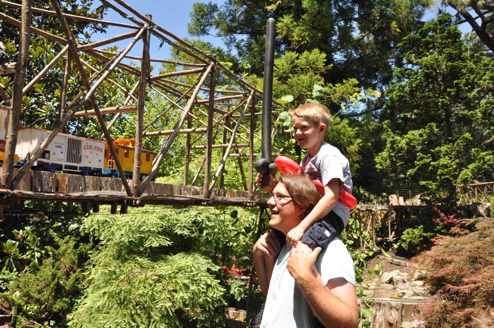 A young boy sits on top of a man's shoulders and watched a model train ride through an miniature elevated bridge.