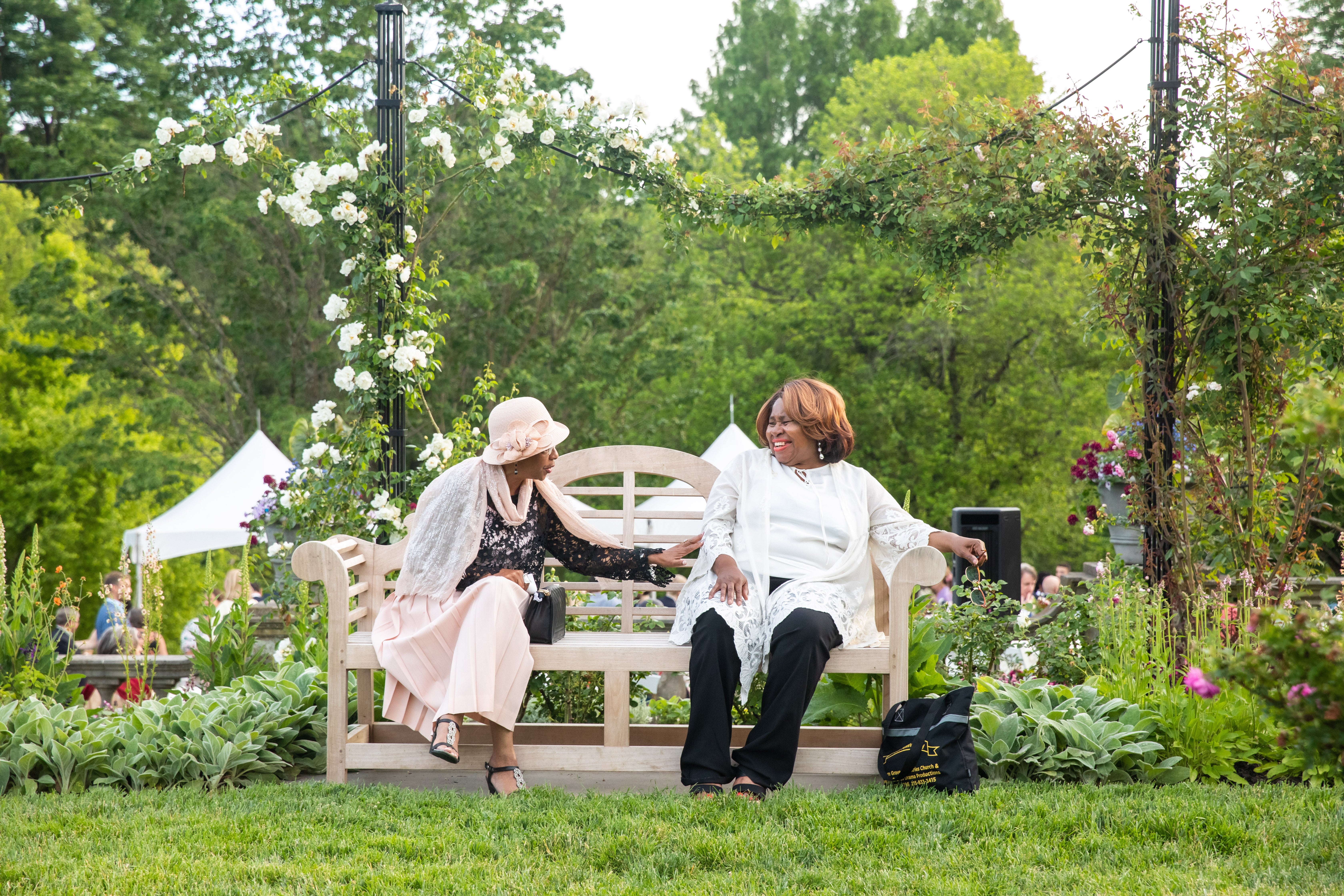 Moonlight&Roses22-142.jpg Two woman talking and laughing on a garden bench surrounded by roses.