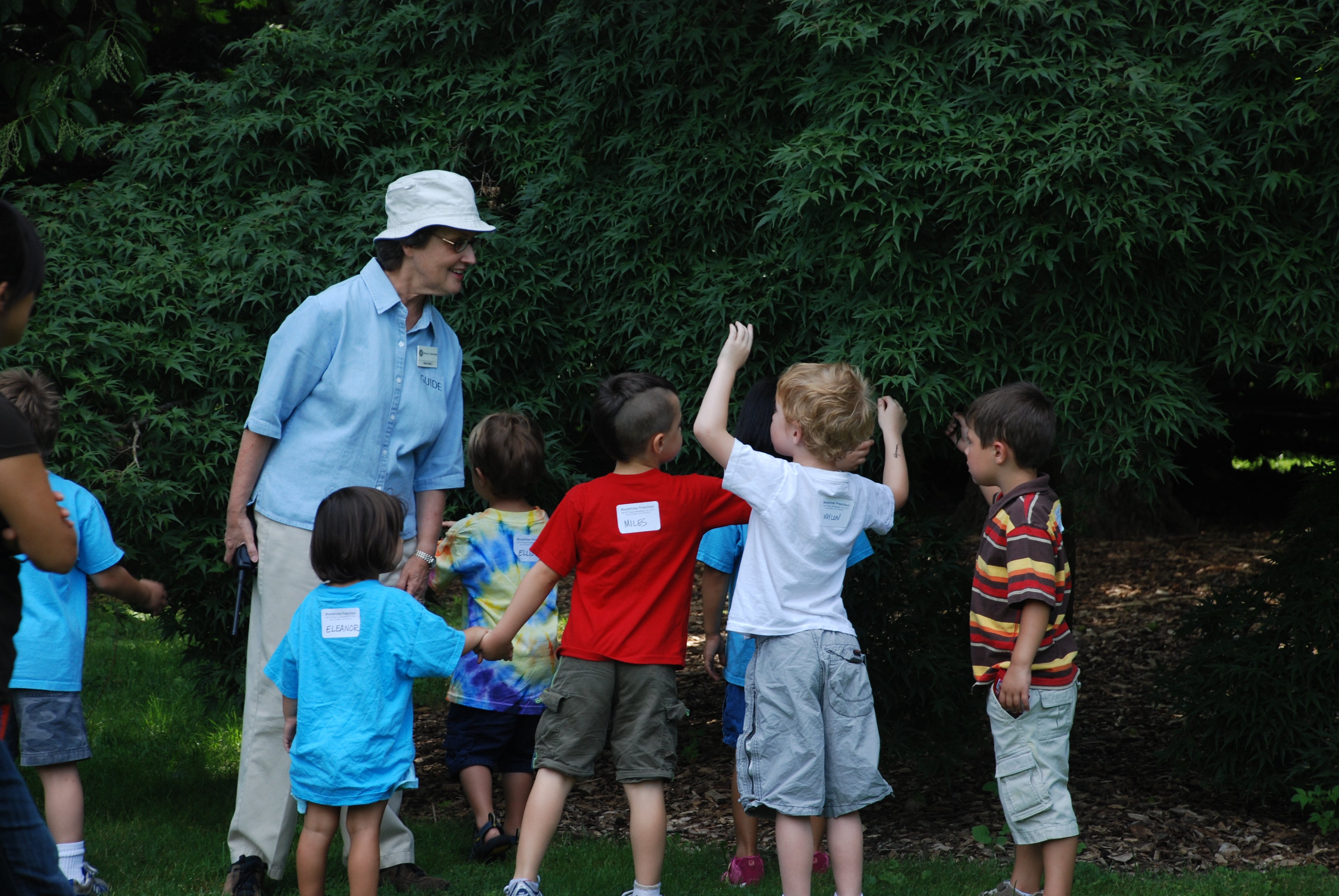 A woman wearing a white bucket hat and blue button up shirt leads a group of children on an outdoors tour.