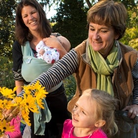 Two woman, one holding a baby in a sling, and a young girl look at yellow flowers smiling. 