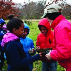 A garden volunteer guides a group of children outdoors in a public garden. 