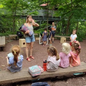 A woman holds her hand up to her ear and holds a drums to a group of young students.