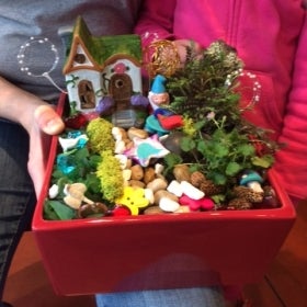 The hands of an adult and child hold a small box filled with plants, miniature gnomes, and fairies. 
