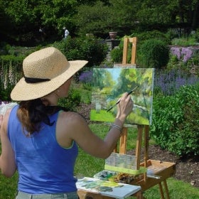 A woman in a hat paints in a garden. 