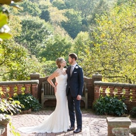 A bride and groom pose in front of a brick railing with trees in the background. 
