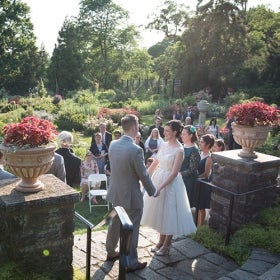 A bride and groom hold hands on a stone entranceway overlooking guests in a rose garden. 