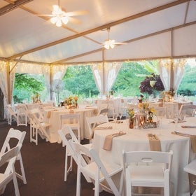 An outdoor wedding tent with round white tables and chairs, flowers, and place settings. 
