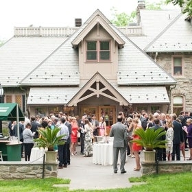 A large group of people mingle outside of a stone building for a wedding cocktail hour. 