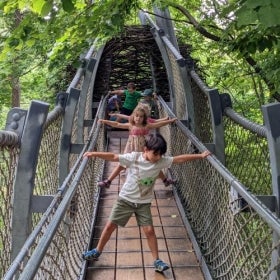 A group of children walk along an elevated bridge in a line with their arms outstretched. 
