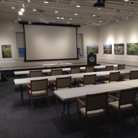 A conference room with white walls, tables, chairs, and a screen. 