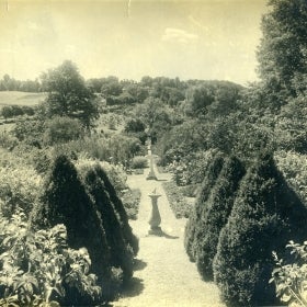 A black and white photo of a garden with a sundial and marble fountain. 