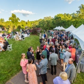 A large group of people mingle outside at white tables and tents.