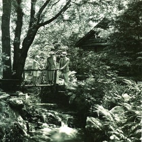 A black and white photo of two adults and a child looking into a stream outside of a log cabin.