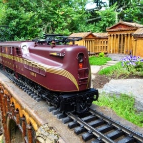 A maroon model train rides along a track next to a miniature replica of the Philadelphia Art Museum. 