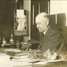 A man writes at a desk covered in botanical material in 1925. 