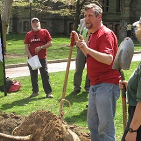 A man in a red shirt holds a shovel in a mound of dirt and speaks to a group of people.