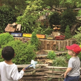 Two young boys watch a model train ride by in an outdoor garden railway.