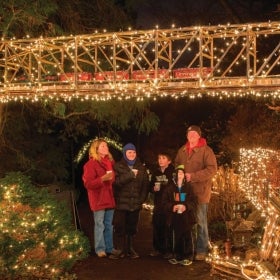A family looks up at a miniature trestle decorated in lights, as part of a model train display.