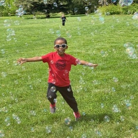 A young boy wearing sunglasses extends his arms to the sides while running through bubbles in a sunny green field. 