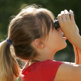 A young girl in a red shirt looks up at the sky with binoculars. 