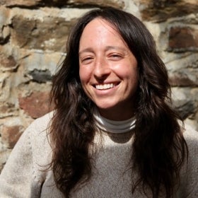 Headshot of a woman with long brown hair wearing a beighe sweater with a white turtleneck underneath in front of a stone wall.