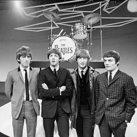 A black and white photograph of The Beatles, all wearing suits and standing in front of a stage with a drum kit and bass drum that reads, "The Beatles."