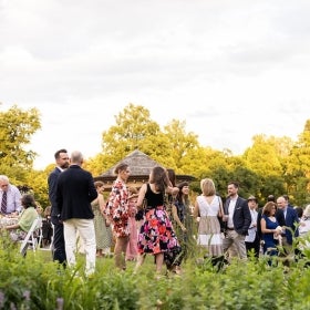 A group of people in informal attire mingle outdoors in a public garden with a gazebo in the background. 