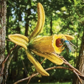 A large fabricated yellow flower with a fabricated bee on top outdoors in a wooded setting.