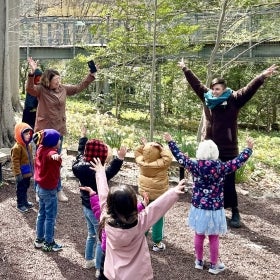 A person leads a group of children and their caregivers with their hands raised and smiling outdoors.