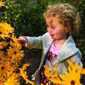 A young girl holds a small magnifying glass while smiling and pointing at a yellow flower.