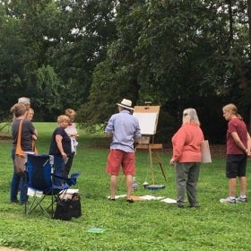 A group of people stand outside around a man with a drawing easel. 