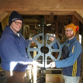 Two men in winter hats hold a large toothed sprocket