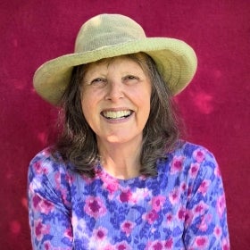 A headshot of a woman with a rimmed hat, purple floral shirt, and a magenta background.