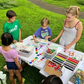 A group of four children stand at a table outdoors water coloring and drawing.