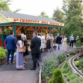A group of people in garden party attire stand outdoors in front of a small yellow, red, and green structure with a sign that reads, "Chestnut Hill Station." 