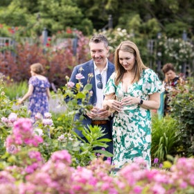 A man and woman in garden party attire stand outdoors on a sunny day in a garden, smiling and admire a bright pink rose bush. 