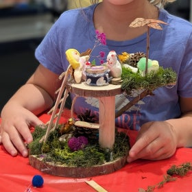 A smiling child posed with a fairyhouse made of natural materials. 