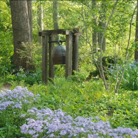 A large sculpture of a bell stand in an outdoor setting surrounded by blue and white flowers and trees. 