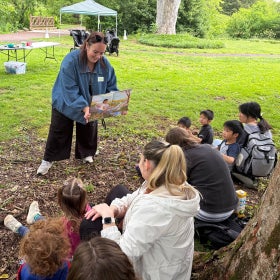 A smiling woman holds up a picture book to a group of children and caretakers in an outdoor setting for story time. 
