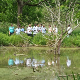 A group of students stand outdoor in a natural wetland area with their reflections mirrored in a pond. 