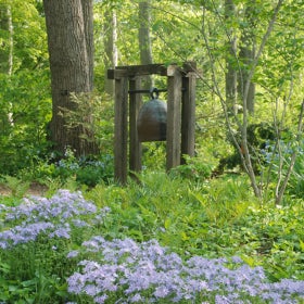 A bronze bell sculpture stands among purple flowers and green foliage. 