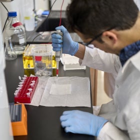 A scientist wearing a lab coat and blue rubber gloves holdings a syringe over a white sheet of paper. 