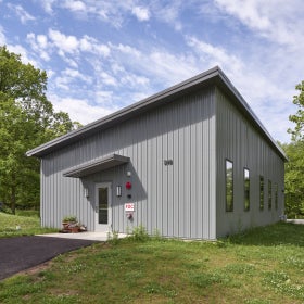 A small grey building with a slanted roof stands in an outdoor green space. 