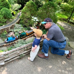 An older man and a young boy look at an outdoor model train display.
