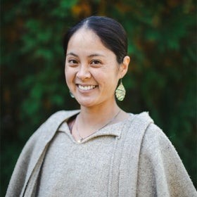 A headshot of a woman smiling at the camera with dark hair pulled back, dangling earrings, and an oatmeal-colored sweater. 