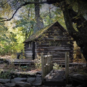 A log cabin with a small bridge leading to it surrounded by trees and fall foliage. 