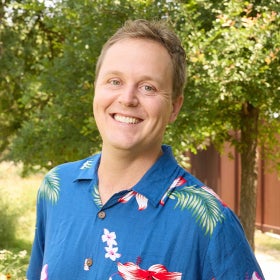 A headshot of a man wearing a bright blue Hawaiian shirt with red and pink flowers. 