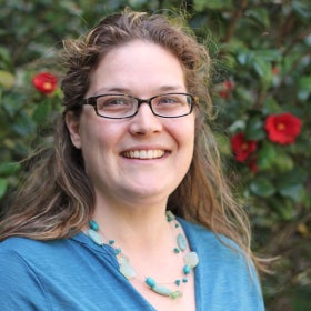 A headshot of a woman wearing a blue shirt and blue beaded necklace in front of a shrub in bloom with red flowers. 