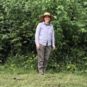 A woman stands in an outdoor green space wearing a wide brimmed hate and smiling. 