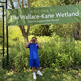 A young boy holds binoculars to his eyes outdoors in front of a sign that reads, "Welcome to The Wallace-Kane Wetland, Open to Explore Year-Round."